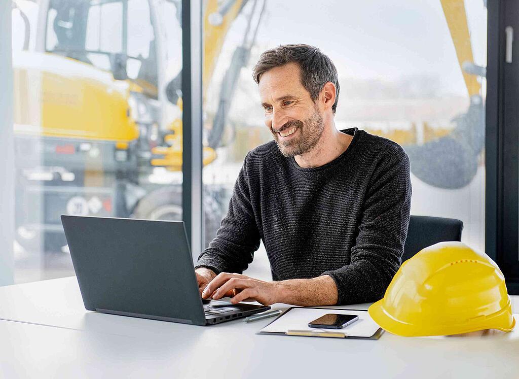 A man works on his laptop next to a yellow Wacker Neuson construction helmet.