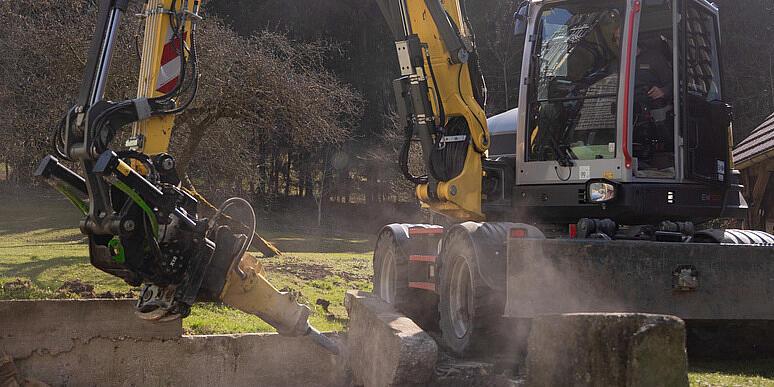 Wacker Neuson EW100 wheeled excavator operating a stump grinder in a forest clearing, grinding a tree stump and throwing wood chips into the air, with stacked logs and woodland in the background.