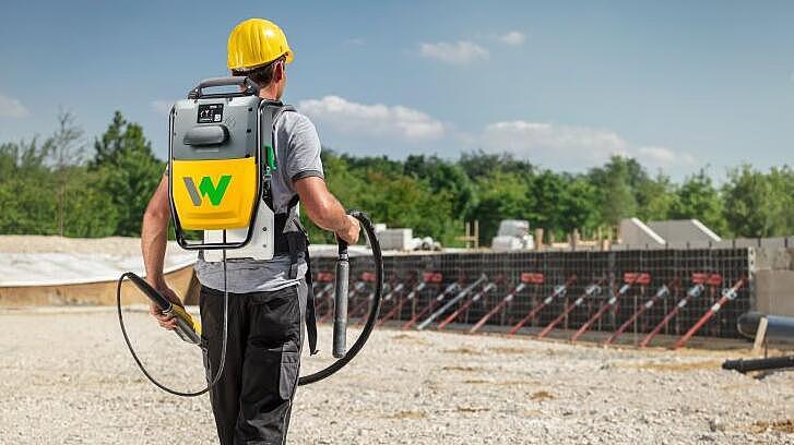 Construction worker carrying battery-powered Wacker Neuson high-frequency internal vibrator on a construction site.