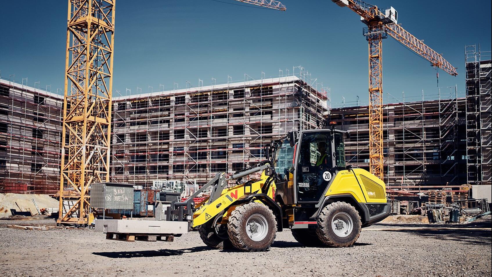 Een Wacker Neuson WL750p-wiellader die betonelementen vervoert over een drukke bouwplaats, met op de achtergrond steigers en torenkranen.