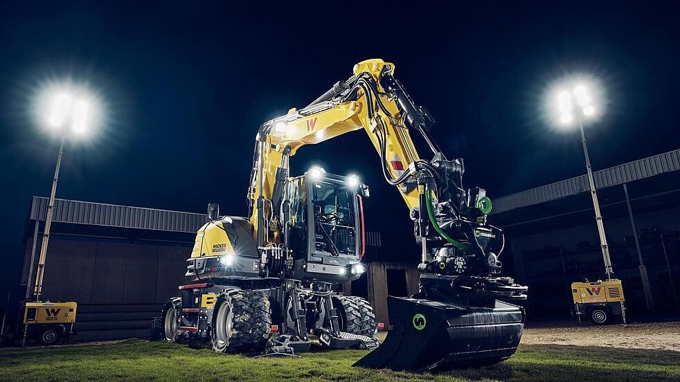 Wacker Neuson EW135-graafmachine op wielen, uitgerust met een tiltrotator en een egalisatiebak, verlicht door schijnwerpers tijdens nachtelijke werkzaamheden.