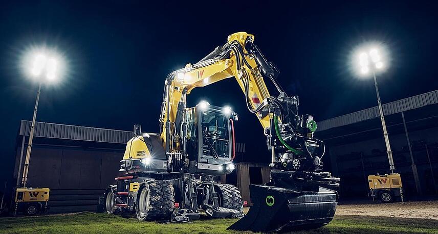Wacker Neuson EW135-graafmachine op wielen, uitgerust met een tiltrotator en een egalisatiebak, verlicht door schijnwerpers tijdens nachtelijke werkzaamheden.