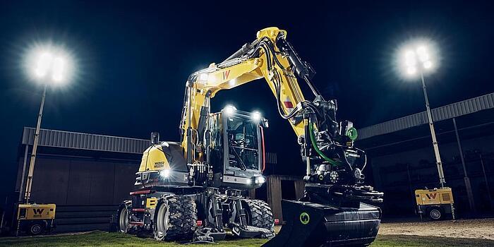 Wacker Neuson EW135-graafmachine op wielen, uitgerust met een tiltrotator en een egalisatiebak, verlicht door schijnwerpers tijdens nachtelijke werkzaamheden.