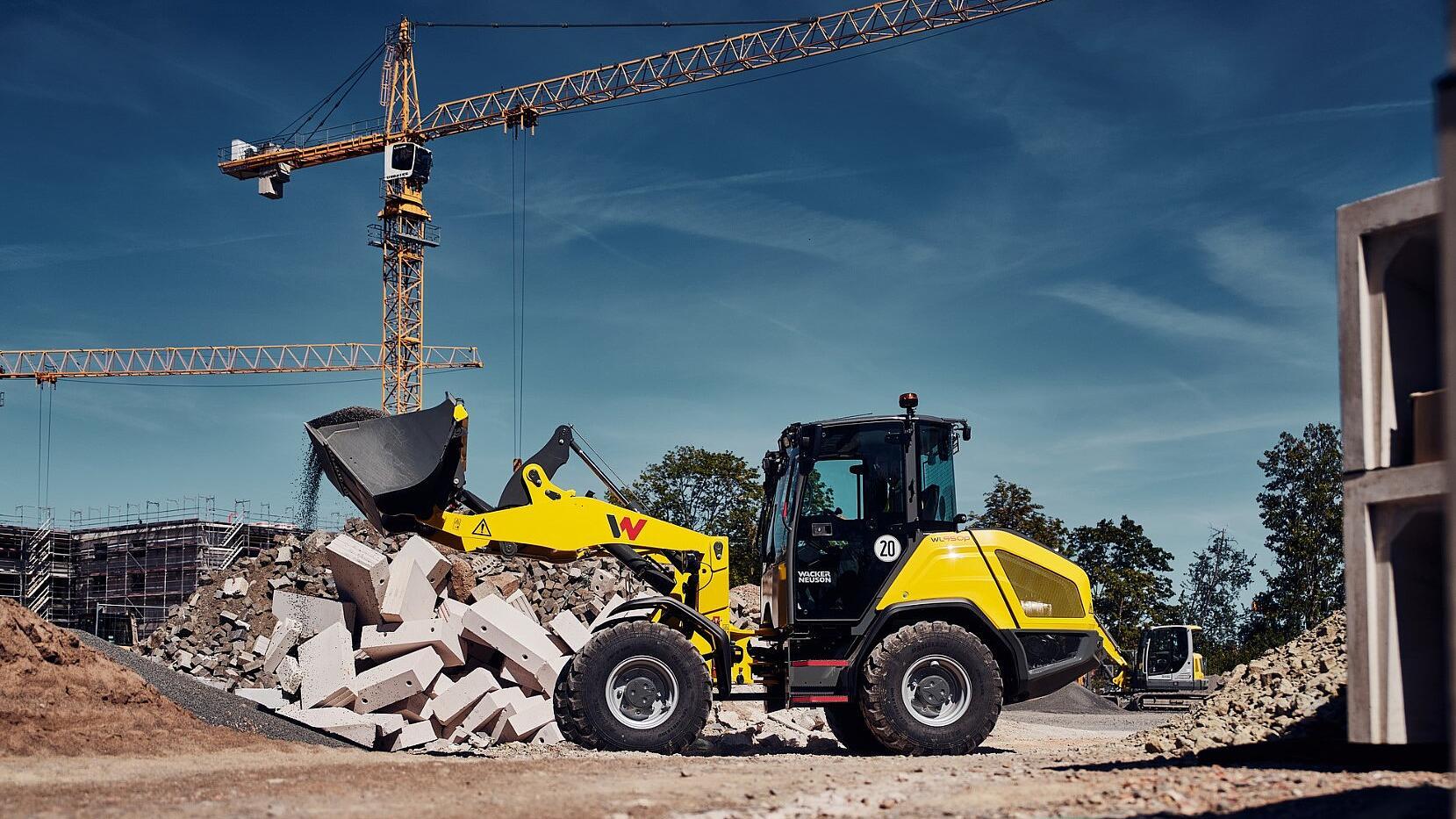 Een Wacker Neuson WL950p-wiellader lost puin uit een laadbak op een stapel sloopafval op een bouwplaats, met torenkranen op de achtergrond.