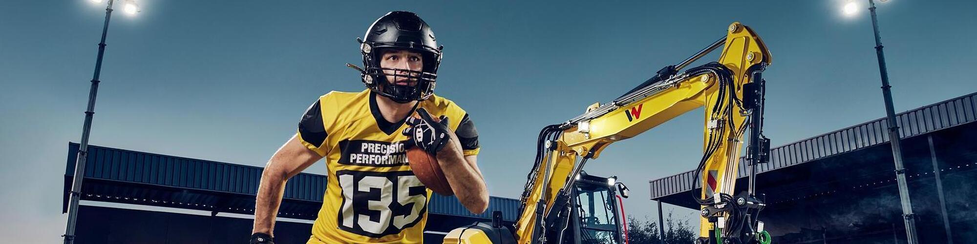 American football player running with a football in the foreground, wearing a yellow jersey with the text “Precision Performance 135”, positioned in front of a yellow Wacker Neuson EW135 wheeled excavator under stadium floodlights.