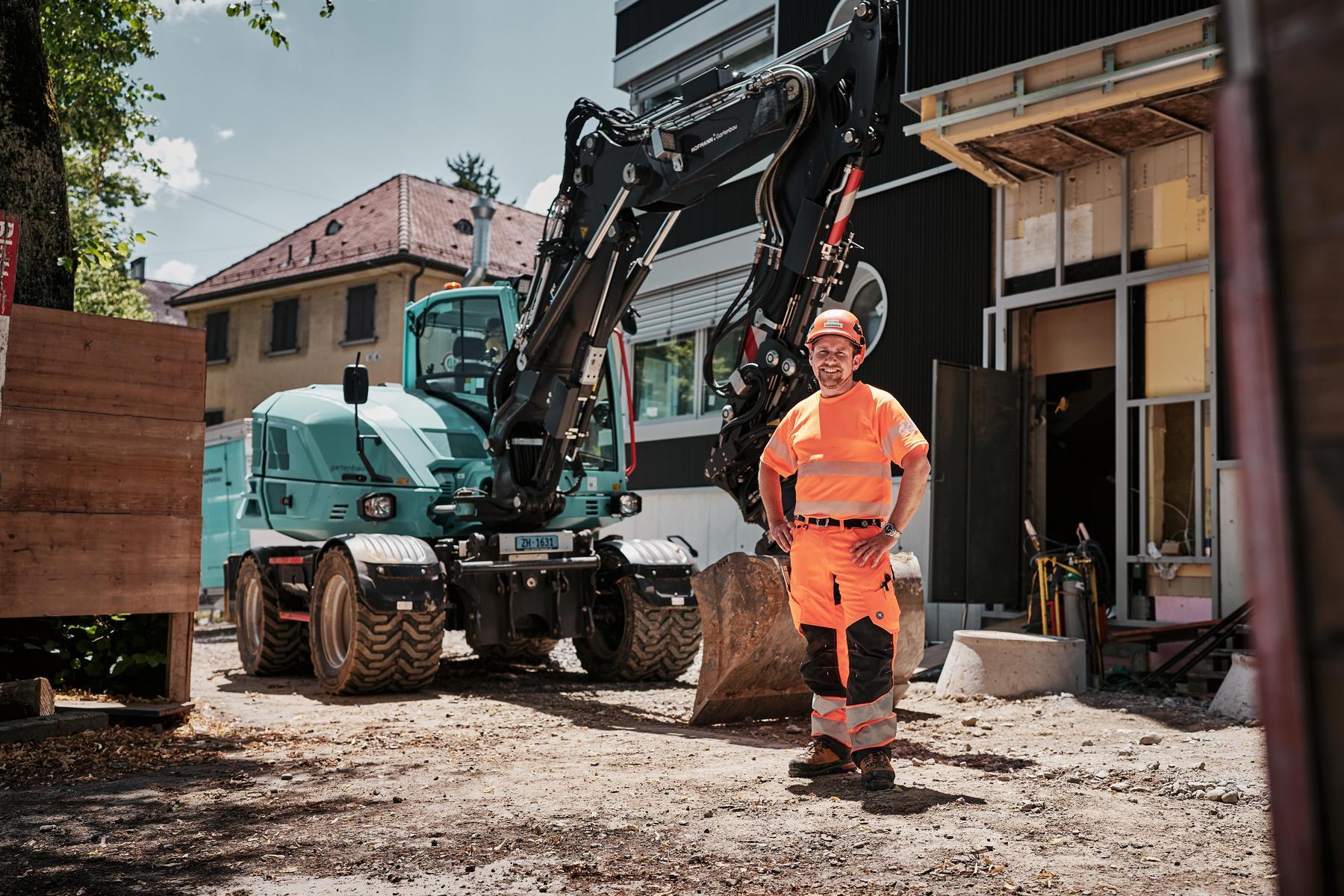 Een machinist in goed zichtbare werkkleding staat naast een Wacker Neuson EW100-graafmachine op wielen op een bouwplaats in de stad.